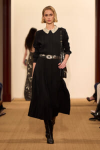 Model on runway wearing a black pleated midi dress with a white lace collar, wide silver belt, knee-high boots, and a black shoulder bag.