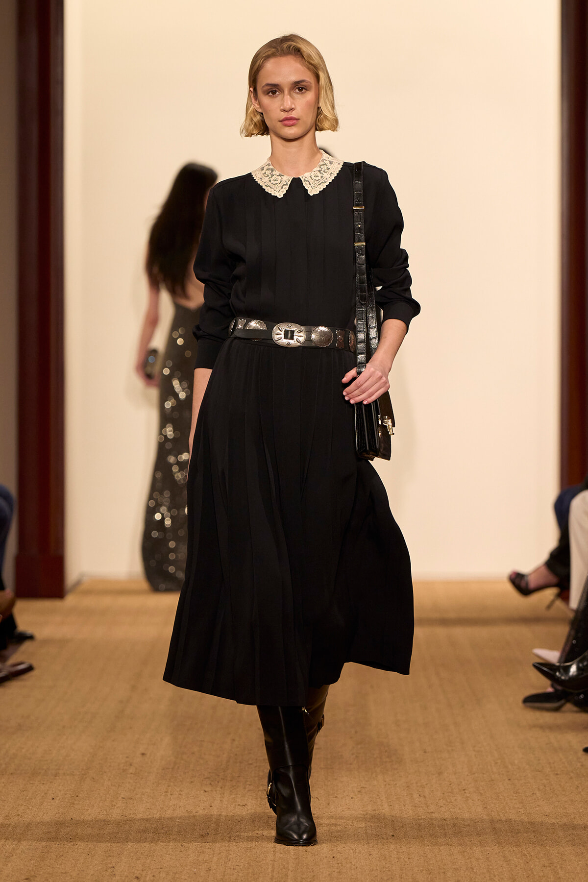 Model on runway wearing a black pleated midi dress with a white lace collar, wide silver belt, knee-high boots, and a black shoulder bag.