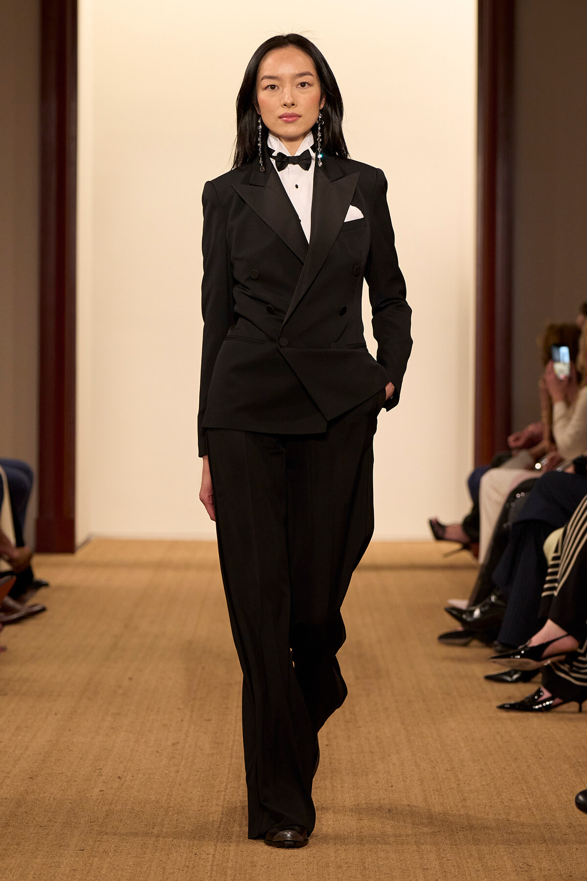 Female model walking a runway in a black double-breasted suit, white shirt, black bow tie, and pocket square.