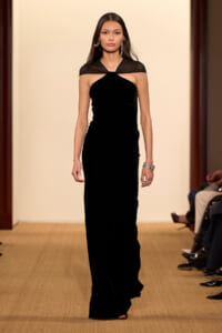 Model walking a runway in a black velvet floor-length gown with a sheer capelet over the shoulders.