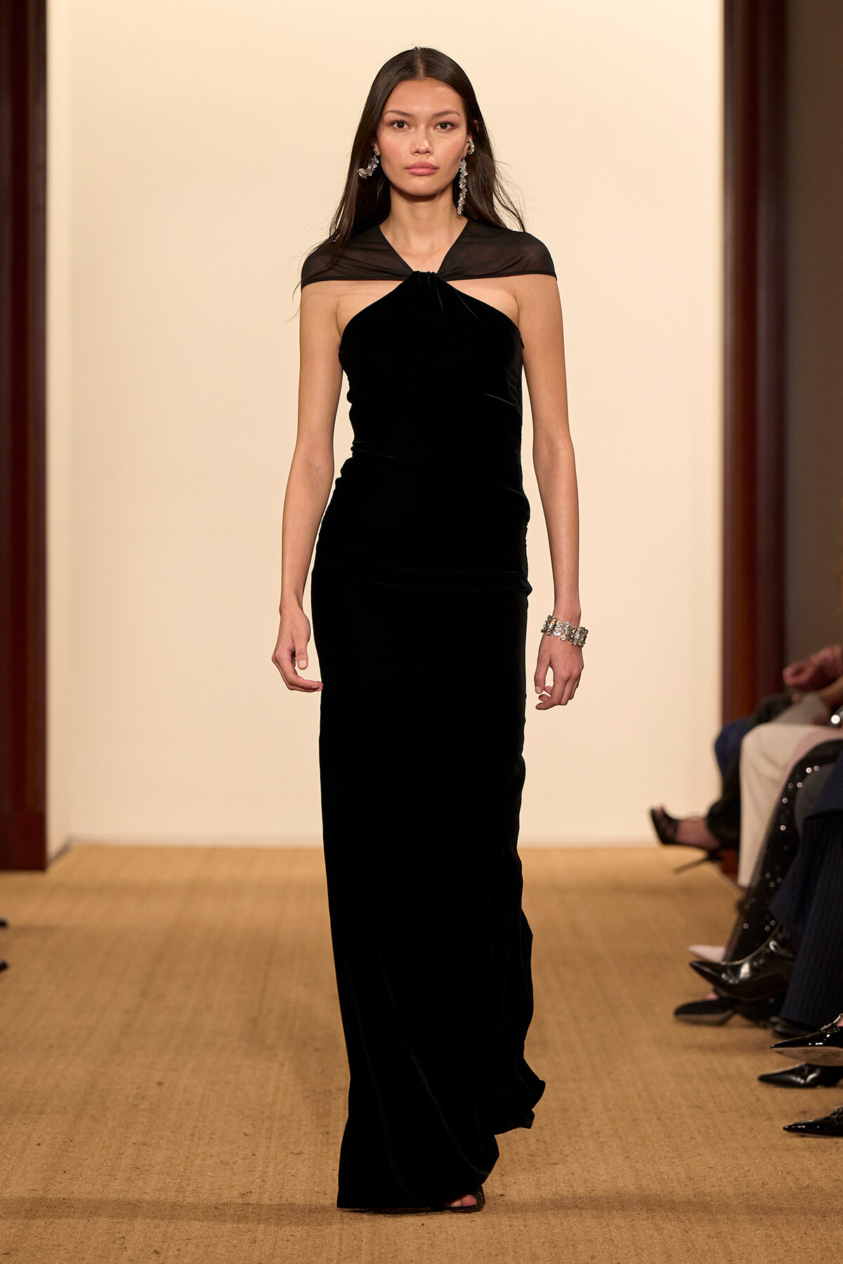 Model walking a runway in a black velvet floor-length gown with a sheer capelet over the shoulders.