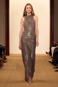 Model walks a runway wearing a sleeveless, high-neck, metallic sequined floor-length gown with dangling earrings against a pale beige backdrop.
