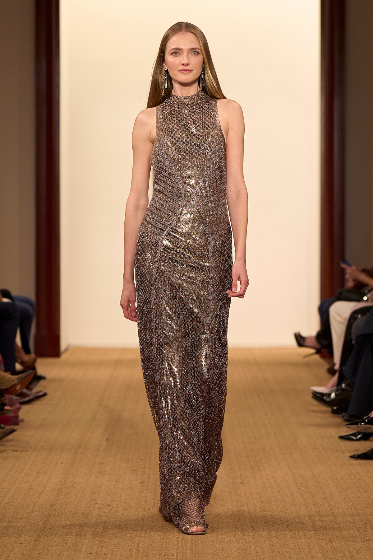 Model walks a runway wearing a sleeveless, high-neck, metallic sequined floor-length gown with dangling earrings against a pale beige backdrop.