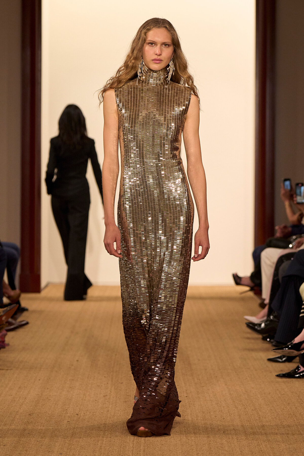 Model walks a runway in a full-length metallic sequined high-neck gown with statement chandelier earrings in a neutral-toned venue.