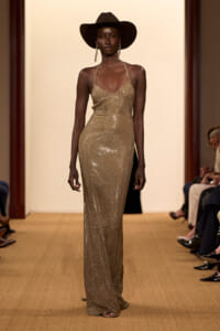 Model walking a runway in a gold sequined halter gown with a wide-brim black hat and dangling earrings.