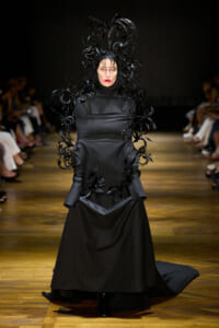 Model walking a runway in a dramatic black gown with a feathered headpiece and cuffs.