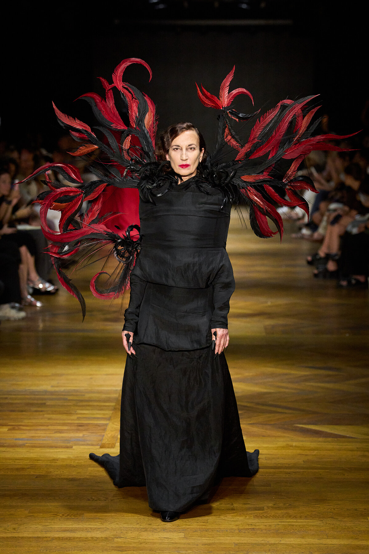 Runway model in a long black gown with large red and black feathered winglike extensions.