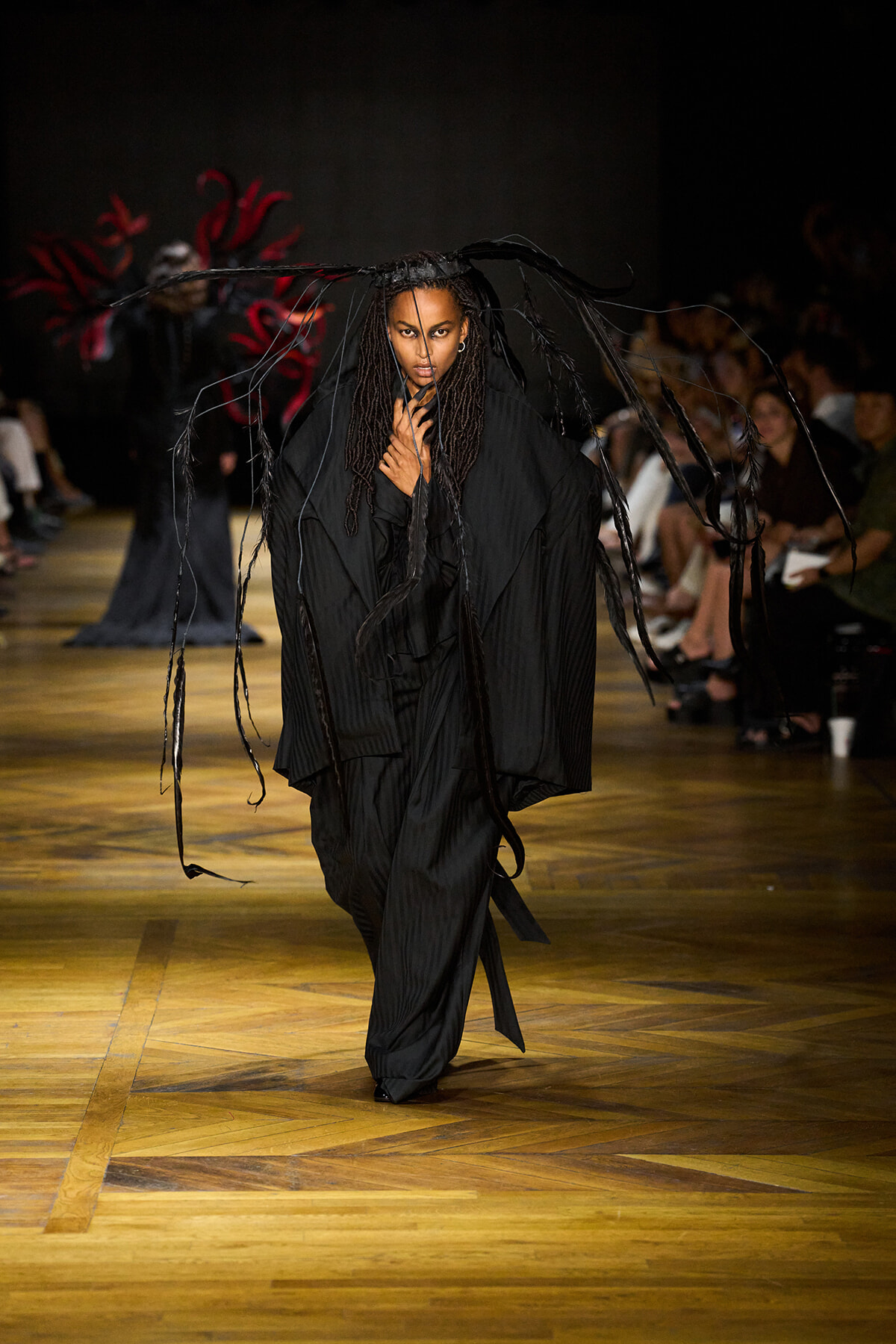 Model in a dramatic black ensemble walks the runway, with long fringe strips hanging around her as the audience watches.