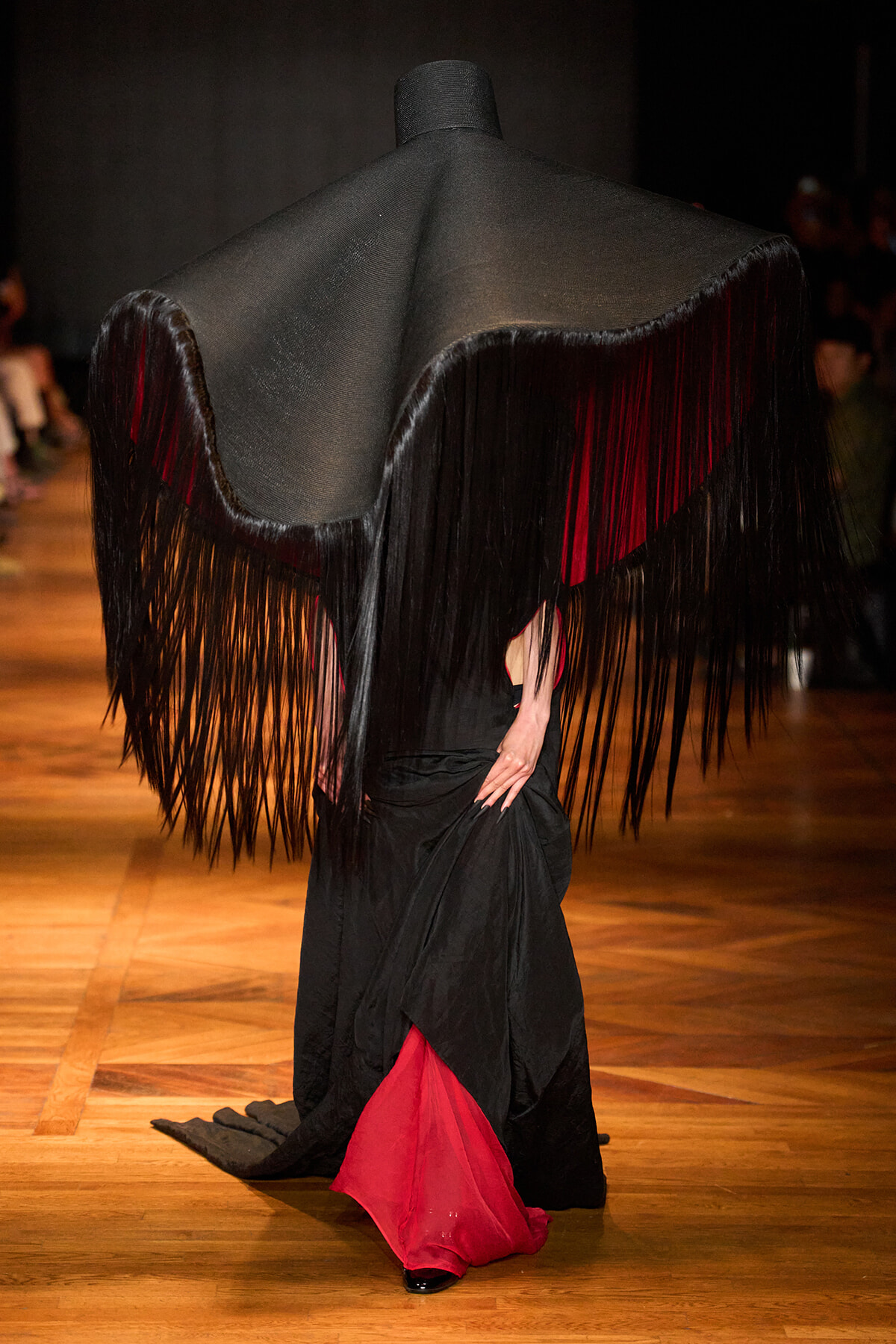 Model on a runway wearing a black cape with long fringe and red lining, standing on a wooden floor.