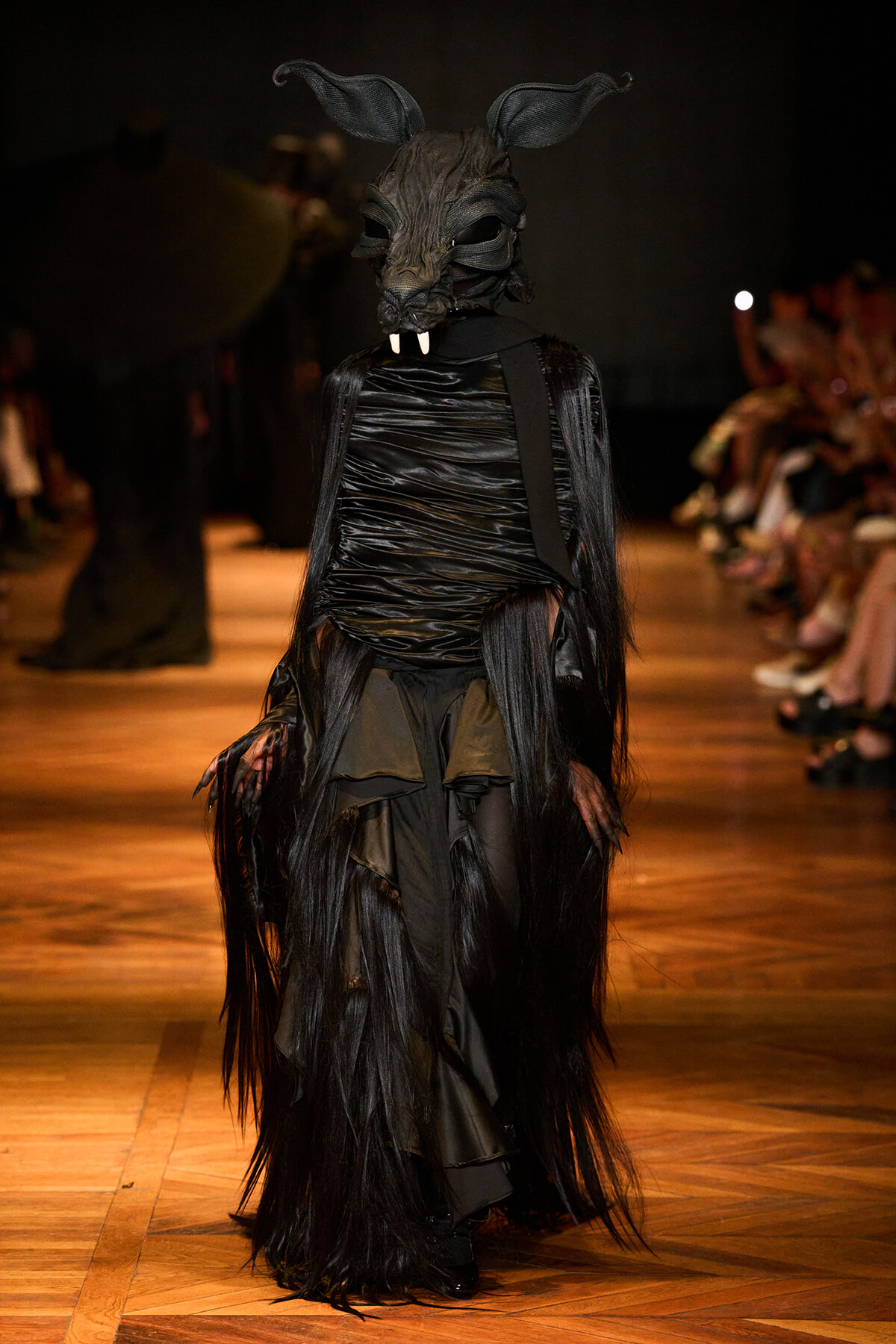 Model on a runway wearing a black dragon-like mask with long fringe and layered dark costume.