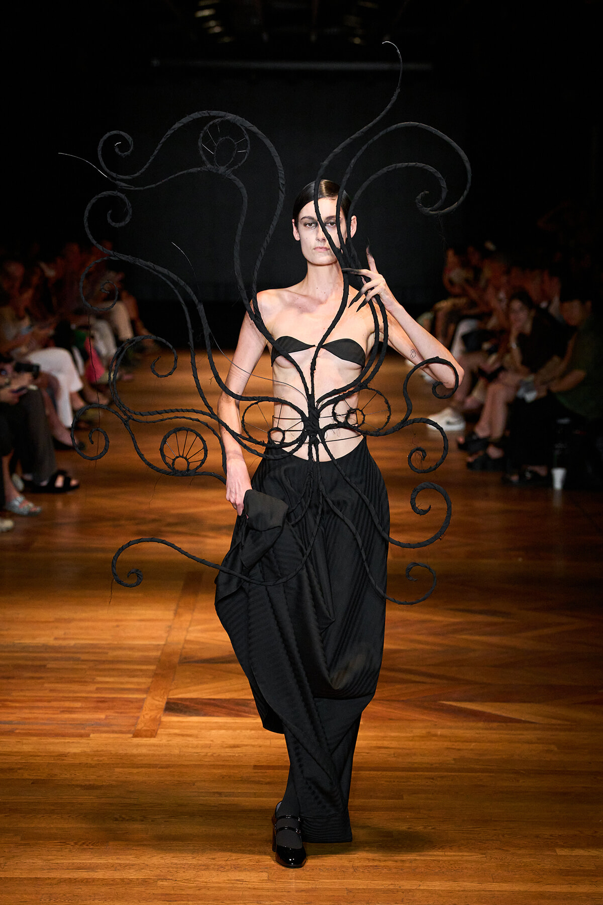 Runway model in a black sculptural gown with long, spiraling wire-like extensions encircling her torso and arms, audience in background.