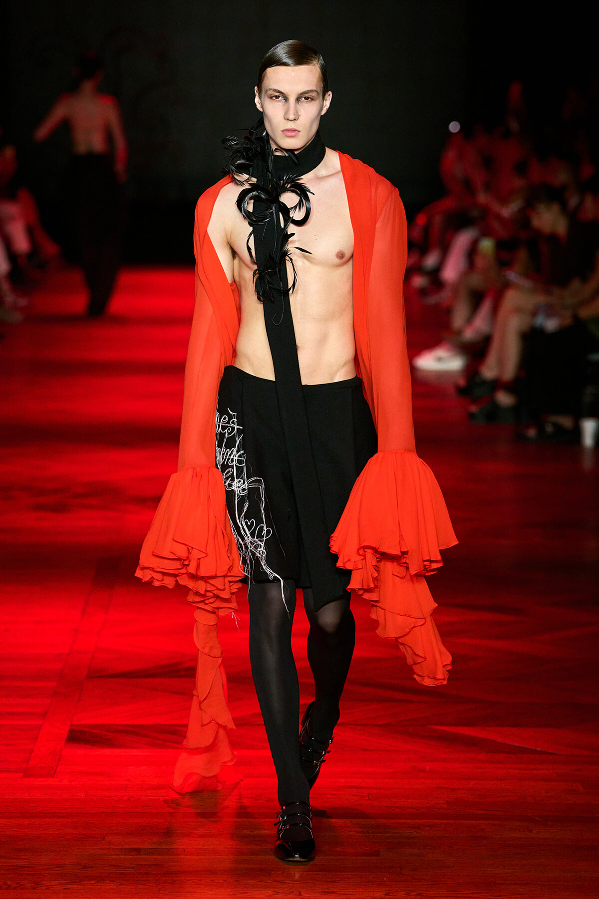 Model on a red-lit runway wearing a red sheer coat with voluminous ruffled sleeves, a black feathered scarf, and a black skirt with white scribble embroidery.