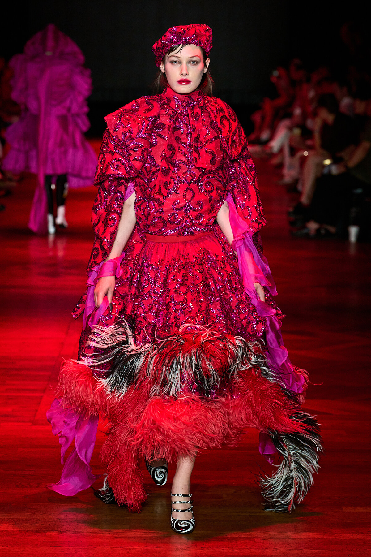 Model walking a runway in a bright red sequined gown with feathered hem and hat, under red lighting