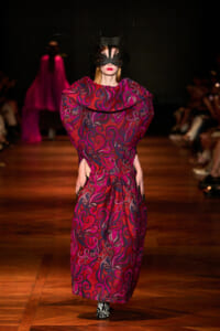 Model on a runway wearing a red-purple swirling-pattern gown with oversized collar and a black feathered mask on the face.