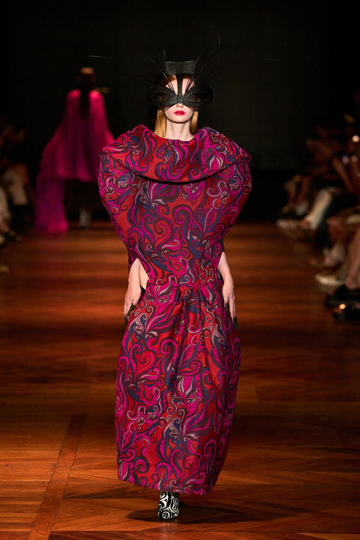 Model on a runway wearing a red-purple swirling-pattern gown with oversized collar and a black feathered mask on the face.