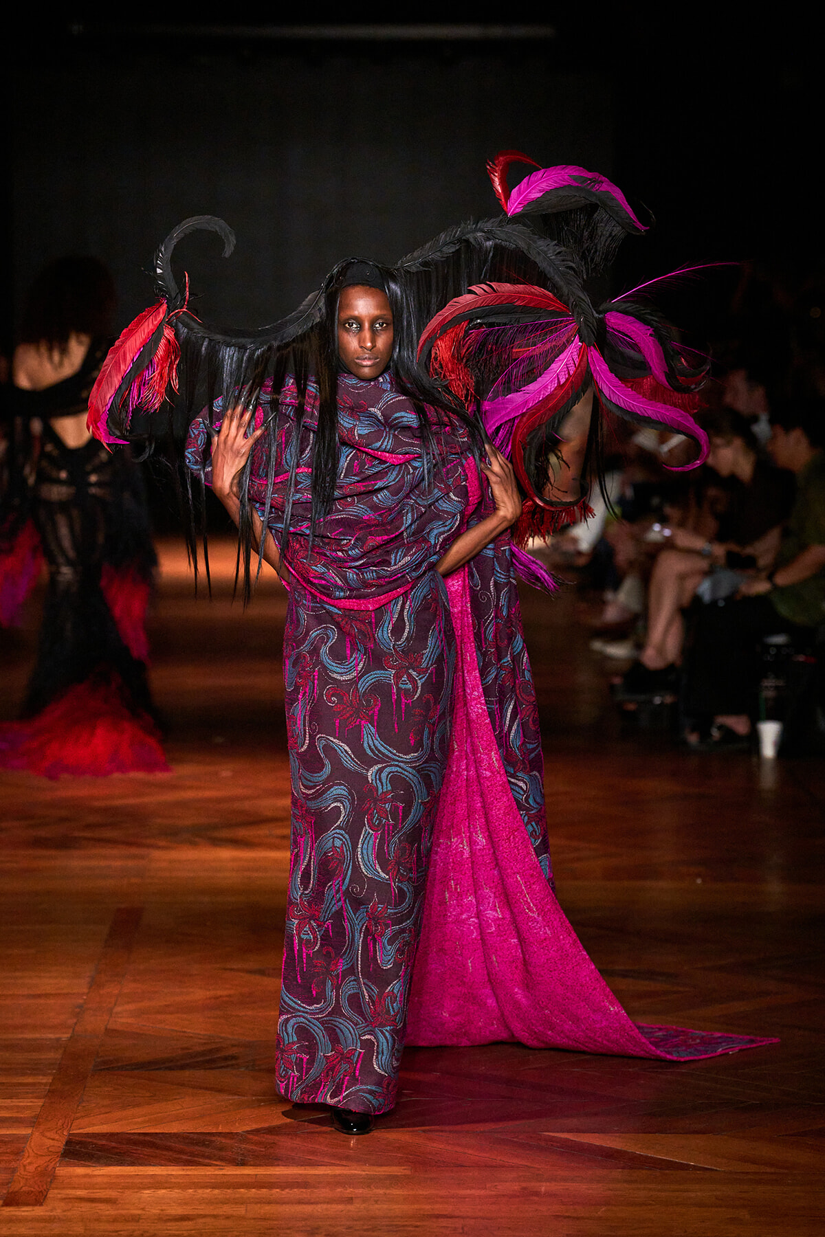 Runway model wearing a long patterned gown with a magenta sash and a dramatic black-and-pink feather headpiece.