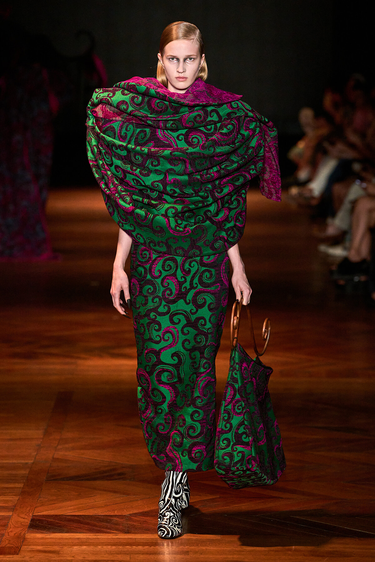 Model walking a fashion runway in a green and magenta swirling pattern dress with a large matching shawl, carrying a patterned bag and wearing black-and-white zebra print boots.