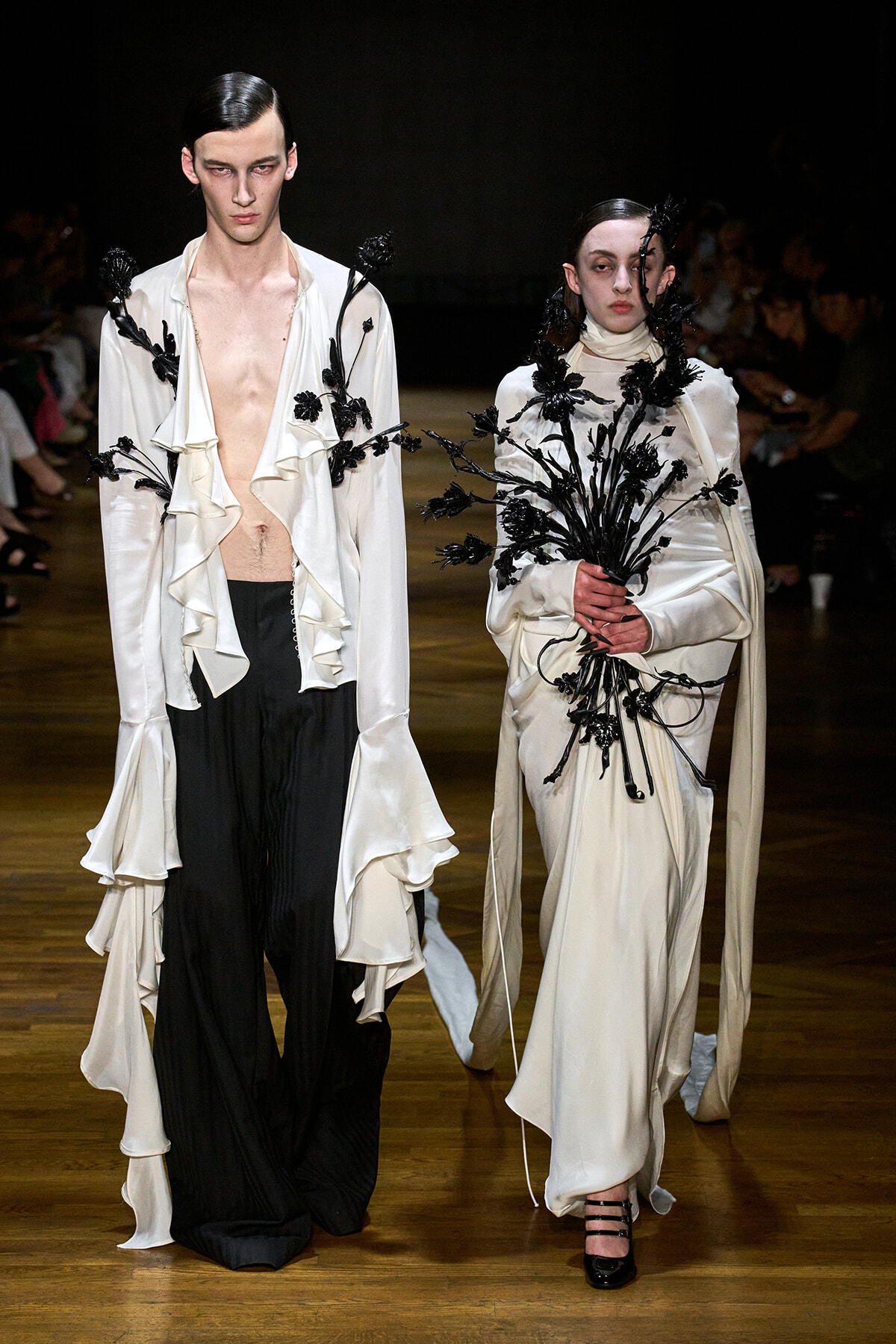 Two models walk a runway in white ensembles with black floral appliqués; left wears a ruffled open-front top with black trousers, right holds a bouquet of black flowers.