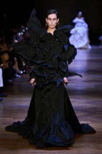 Model walks the runway in a voluminous black dress with sculpted ruffles and a long train, dark backstage lighting setting the mood.