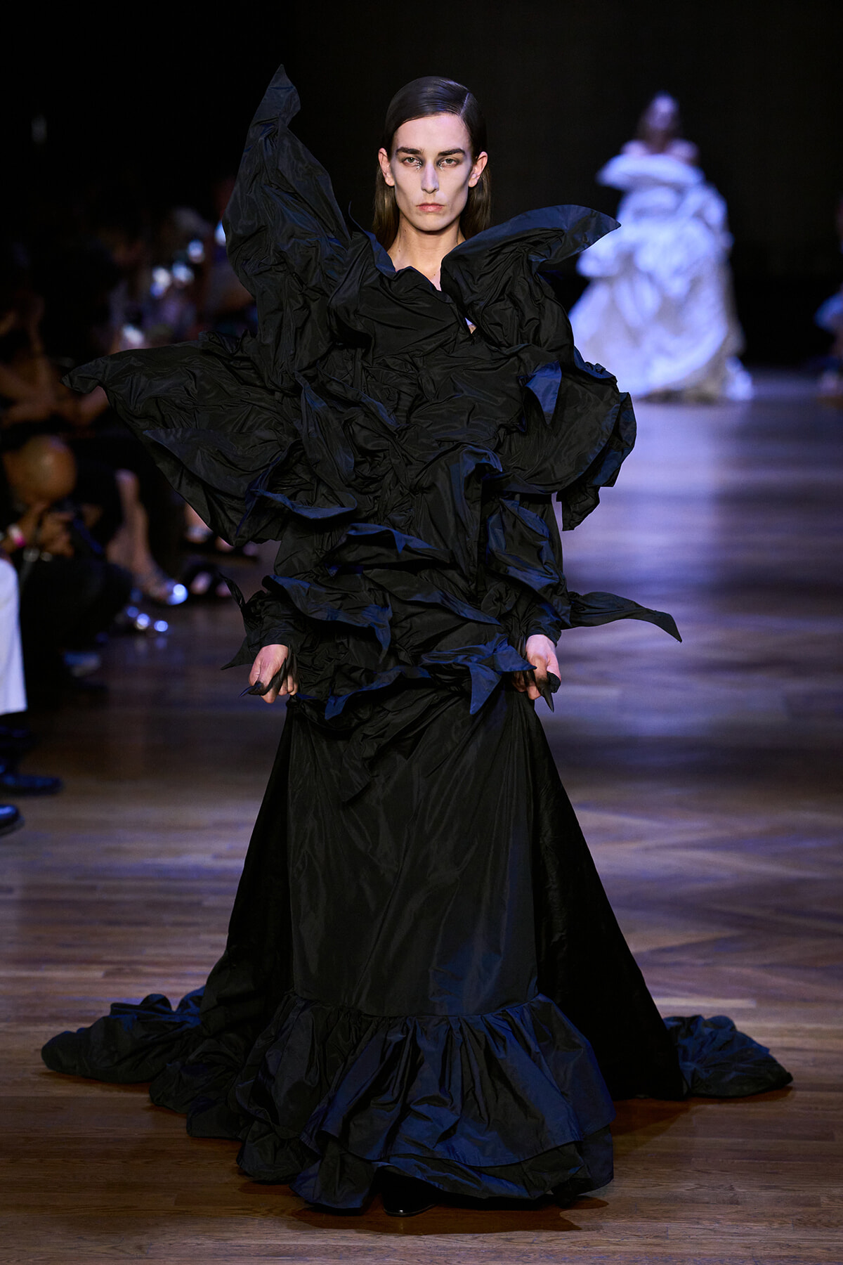Model walks the runway in a voluminous black dress with sculpted ruffles and a long train, dark backstage lighting setting the mood.