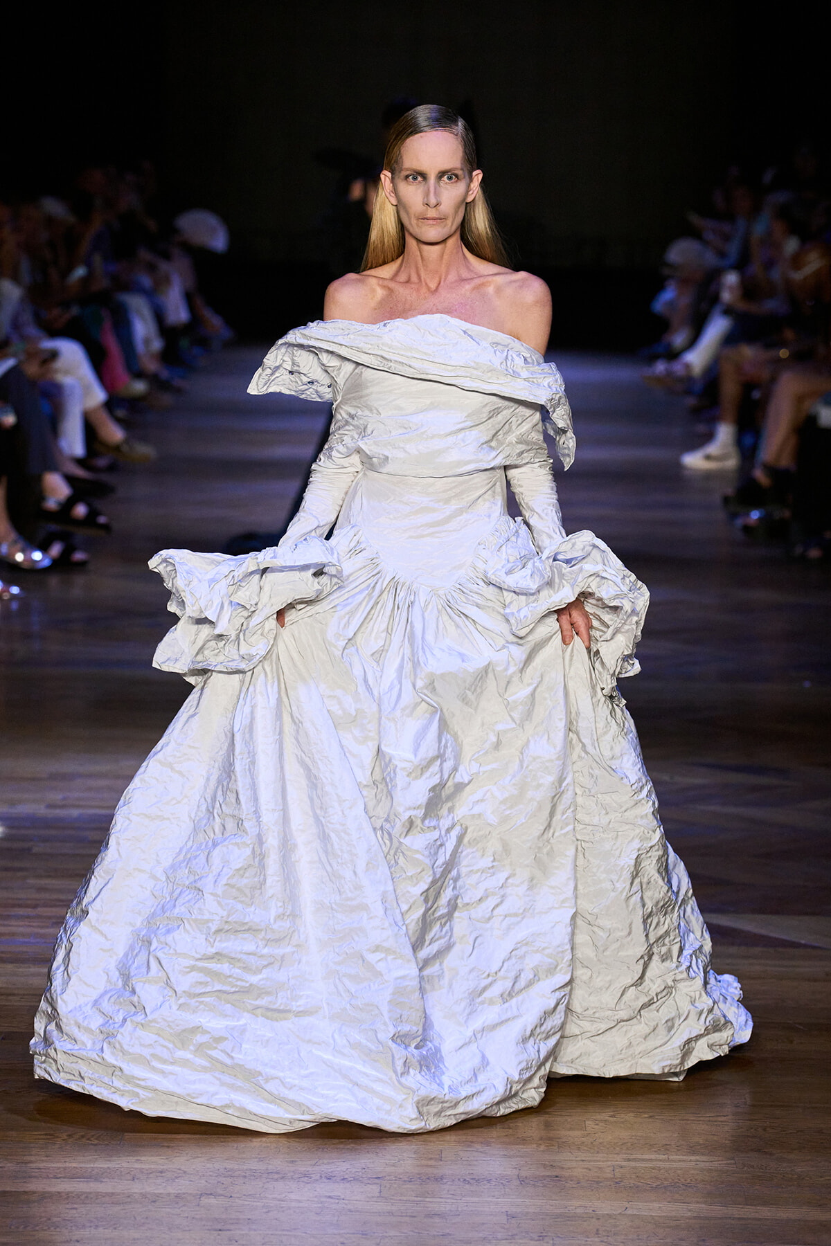 Model wearing a voluminous off-shoulder silver crinkled gown on a runway show.