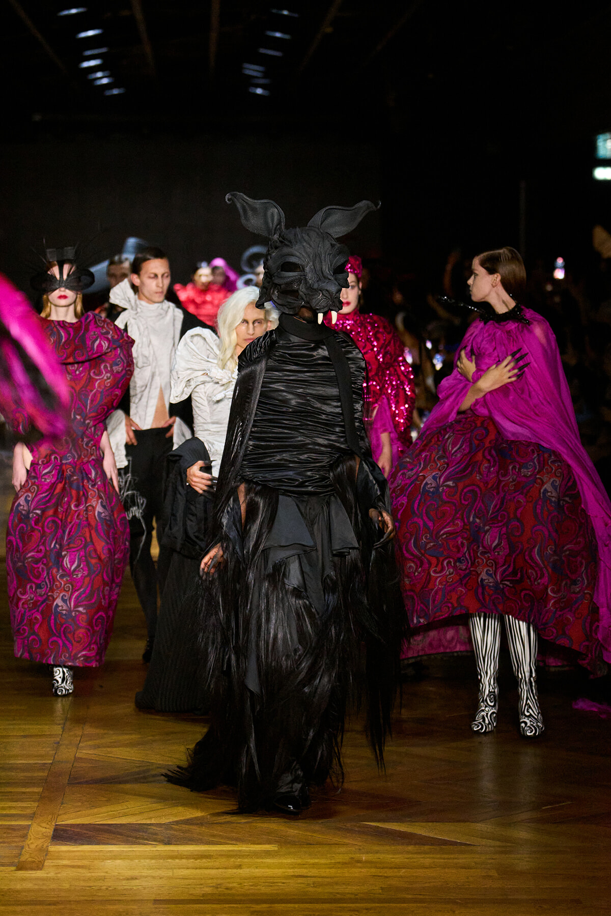 Fashion runway with models in elaborate costumes; central figure wears a black dragon mask and flowing black attire amid purple gowns.