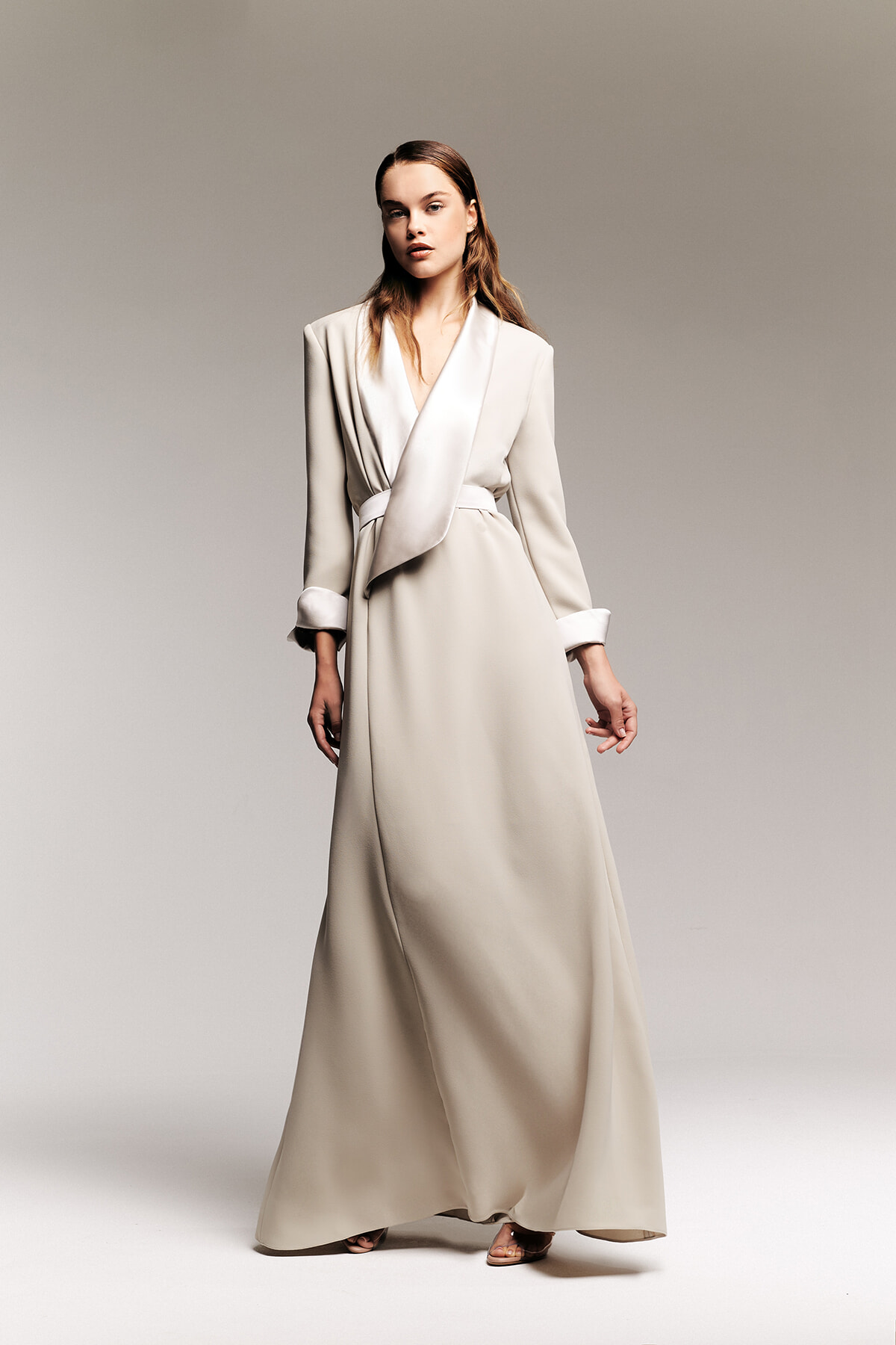 Model wearing a long cream wrap coat dress with a belt, standing against a neutral backdrop.