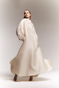 Model wearing a long champagne-colored gown with lace sleeves, posing as the dress flows around her.