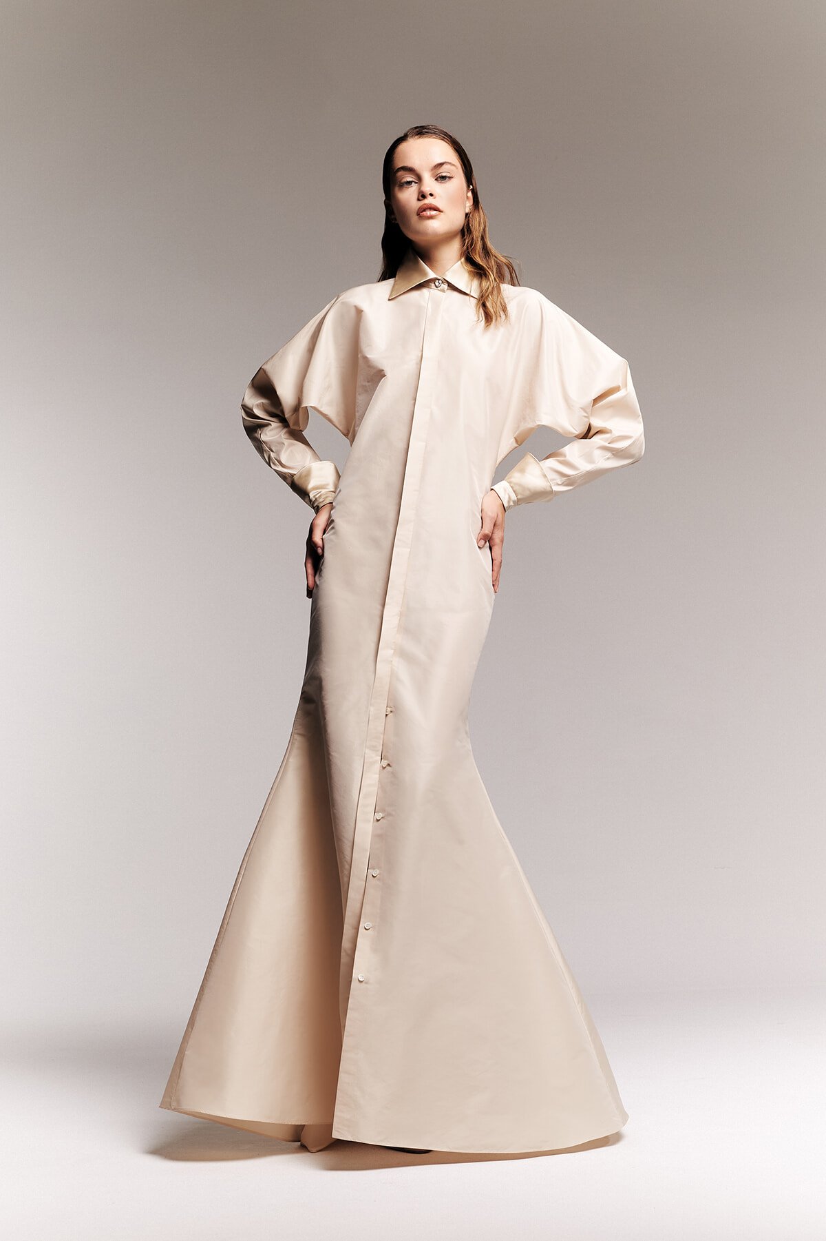 Model wearing a loose, beige button-down maxi dress with oversized sleeves, standing with hands on hips in a studio.