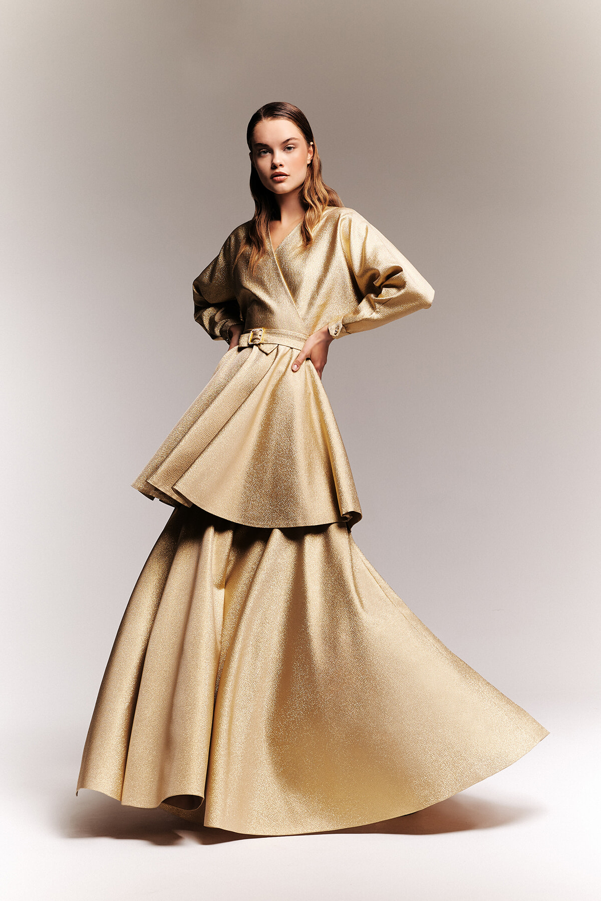 Model wearing a shimmering gold wrap gown with a belted waist and voluminous sleeves, striking a confident pose against a neutral backdrop.