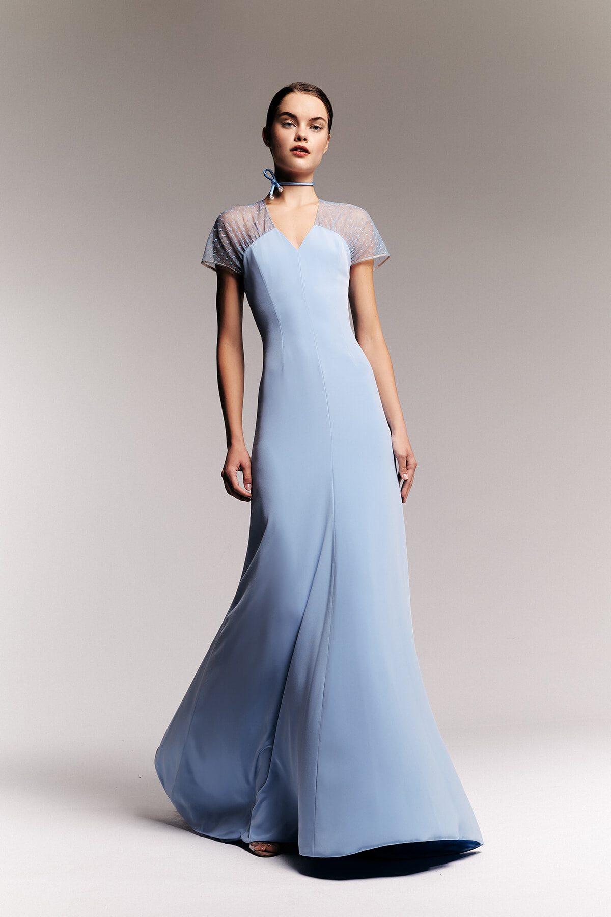Model wearing a pale blue, floor-length satin gown with sheer dotted short sleeves and a matching choker, posing against a neutral backdrop.