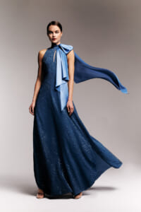 Model wearing a navy blue sequined gown with a light-blue satin scarf tied at the neck and flowing behind her.