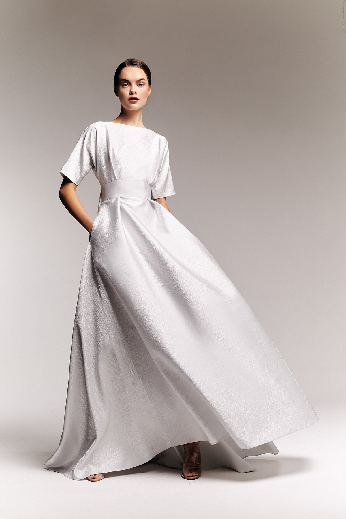 Model wearing a floor-length silver satin gown with a wide waist sash, posing against a neutral backdrop in a studio setting.