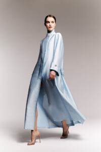 Model in a flowing light blue metallic coat with a high collar, walking and looking to the side in a neutral studio background.