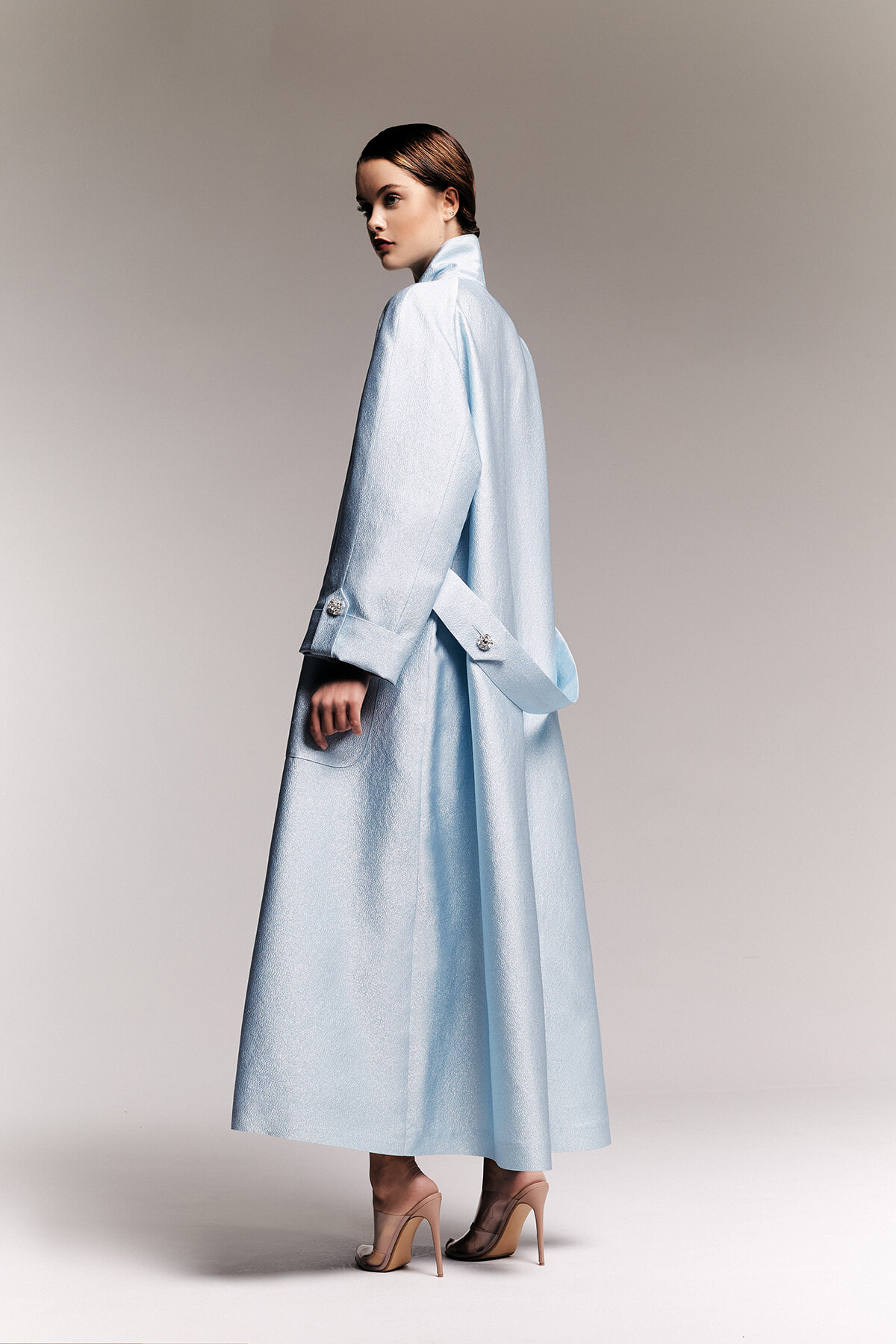 Side view of a woman wearing a light blue, long coat with a back belt, paired with nude high heels.