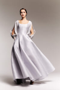 Model wearing a long silver satin gown with dramatic flowing sleeves as she poses against a neutral backdrop