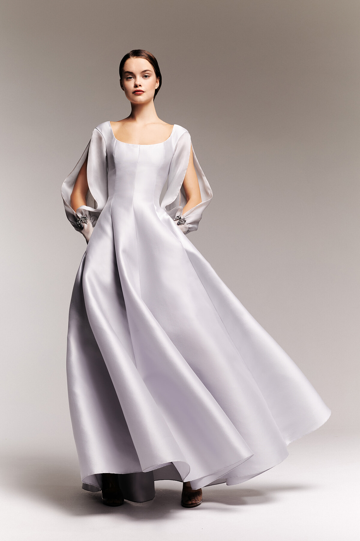 Model wearing a long silver satin gown with dramatic flowing sleeves as she poses against a neutral backdrop