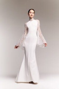 White satin wedding gown with a high lace collar and voluminous lace bell sleeves, modeled standing in a neutral studio backdrop.