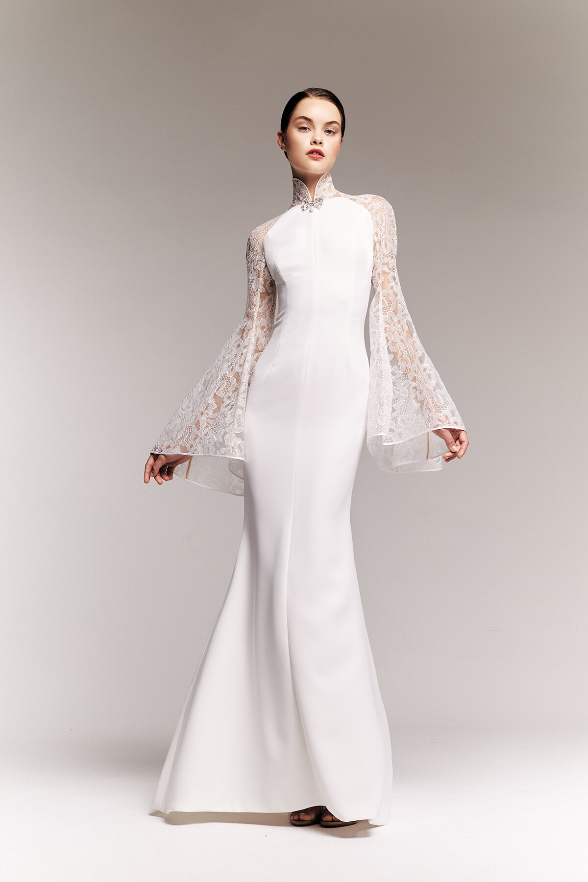 White satin wedding gown with a high lace collar and voluminous lace bell sleeves, modeled standing in a neutral studio backdrop.