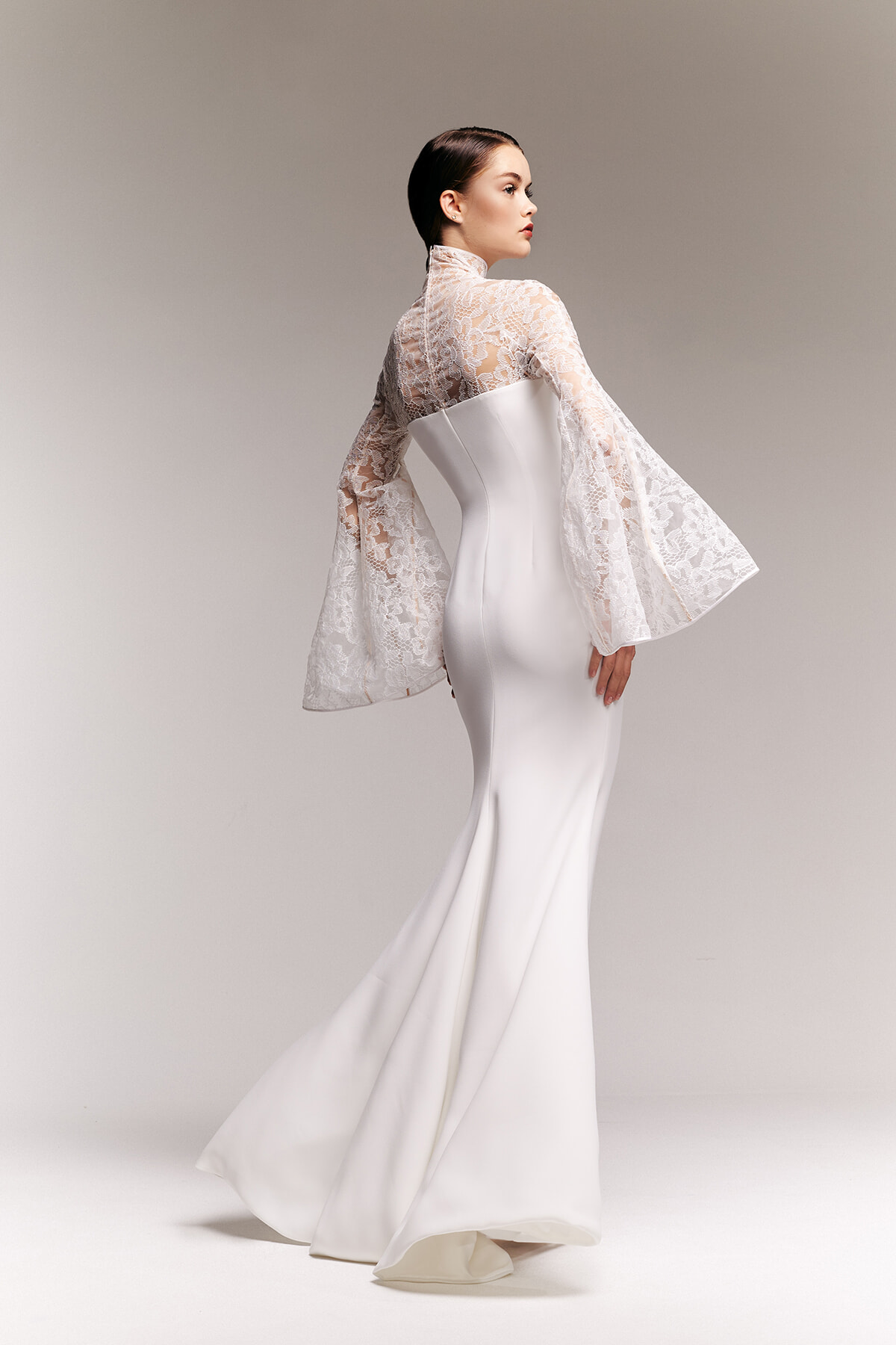 Bride-model in a fitted white bridal gown with a high lace neckline and sheer lace bell sleeves, flowing train behind her.