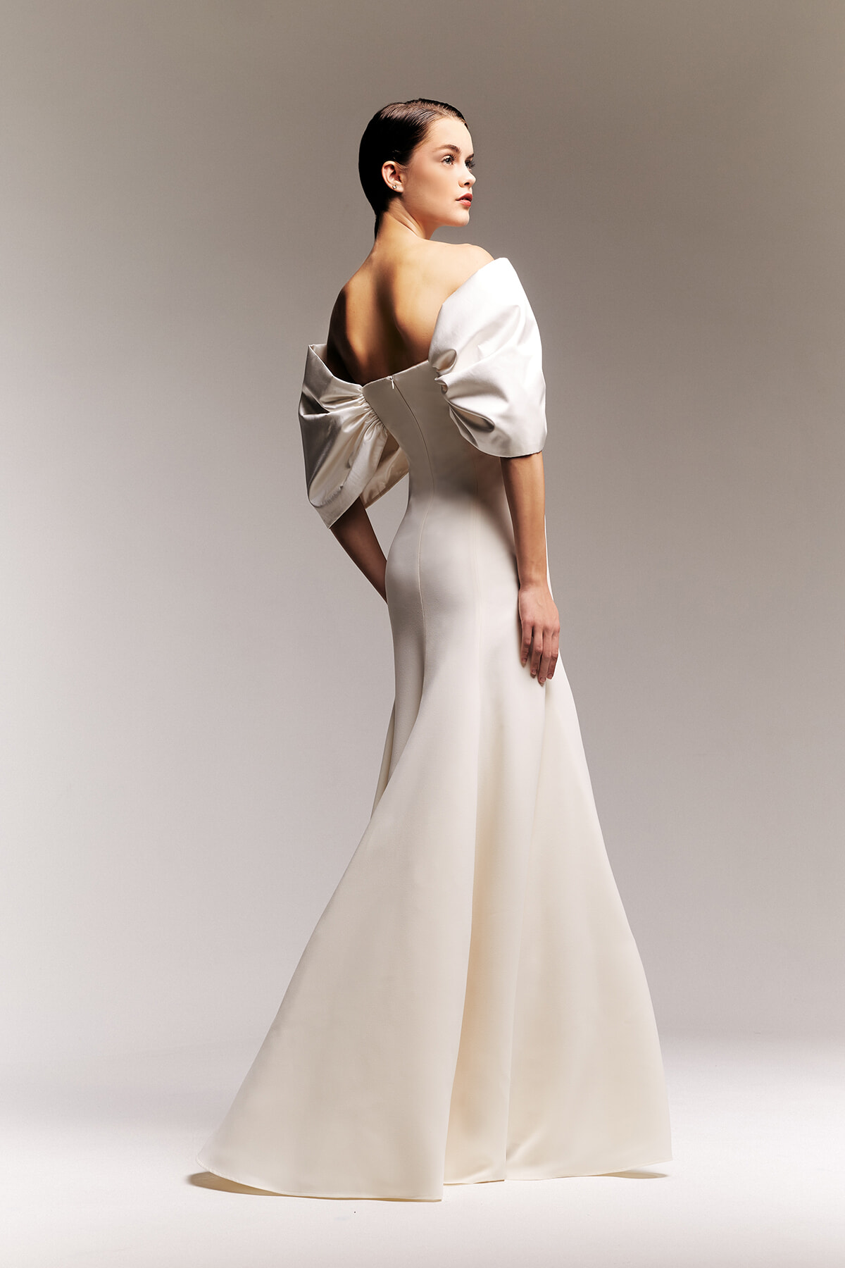 Model wearing an ivory wedding gown with dramatic off-the-shoulder sleeves, shown in profile from the back.