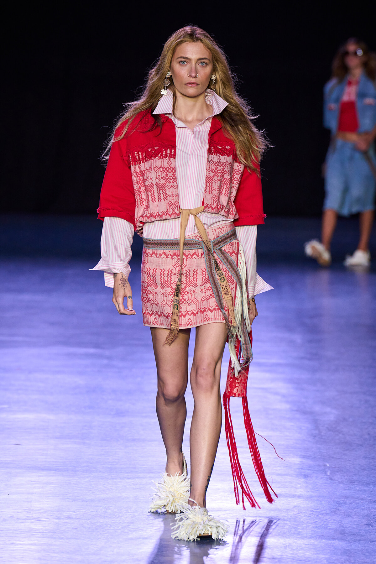 Model walking a fashion runway in a red patterned jacket over a pink striped blouse and matching mini skirt with a woven belt and fringe details.