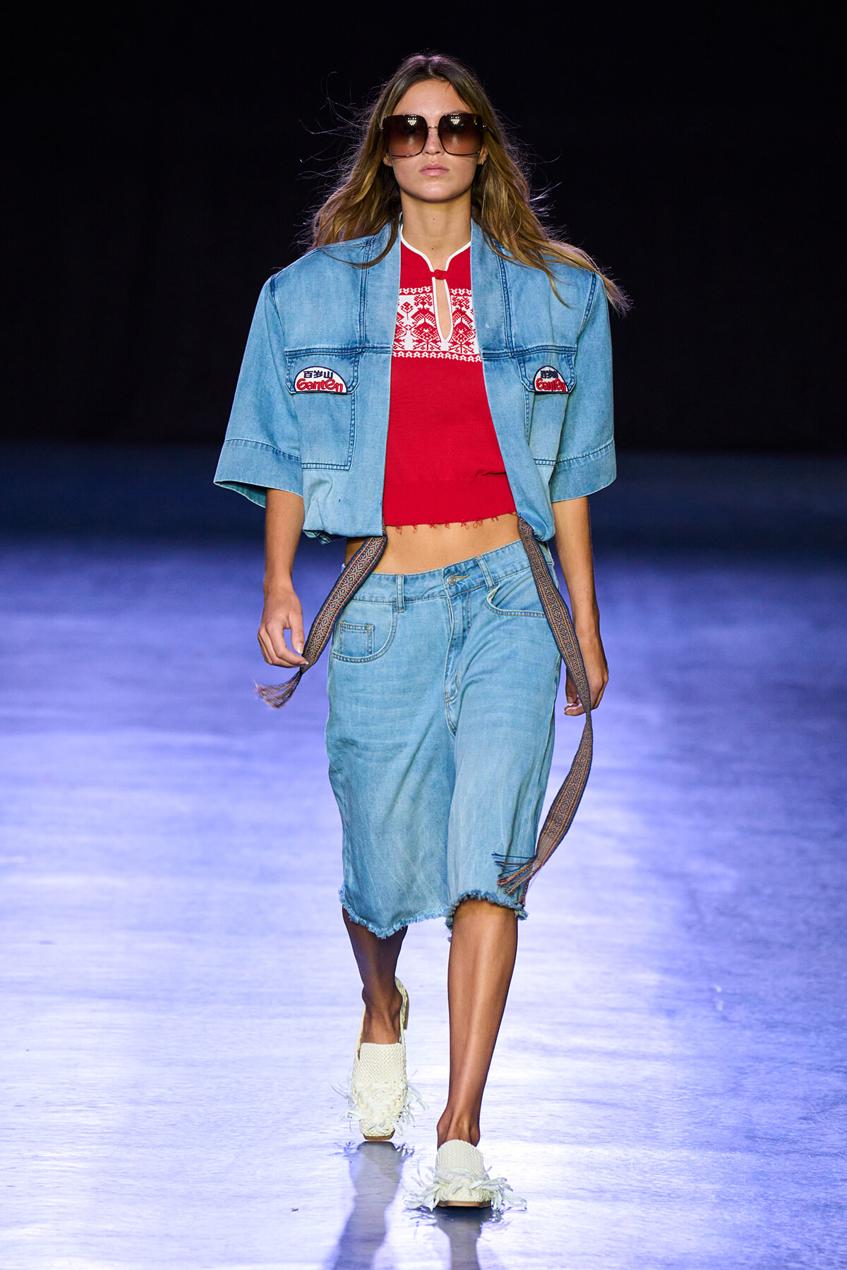 Model walks a runway in a light blue denim set: cropped jacket, red embroidered top, frayed knee-length shorts, and a woven belt with large sunglasses on.