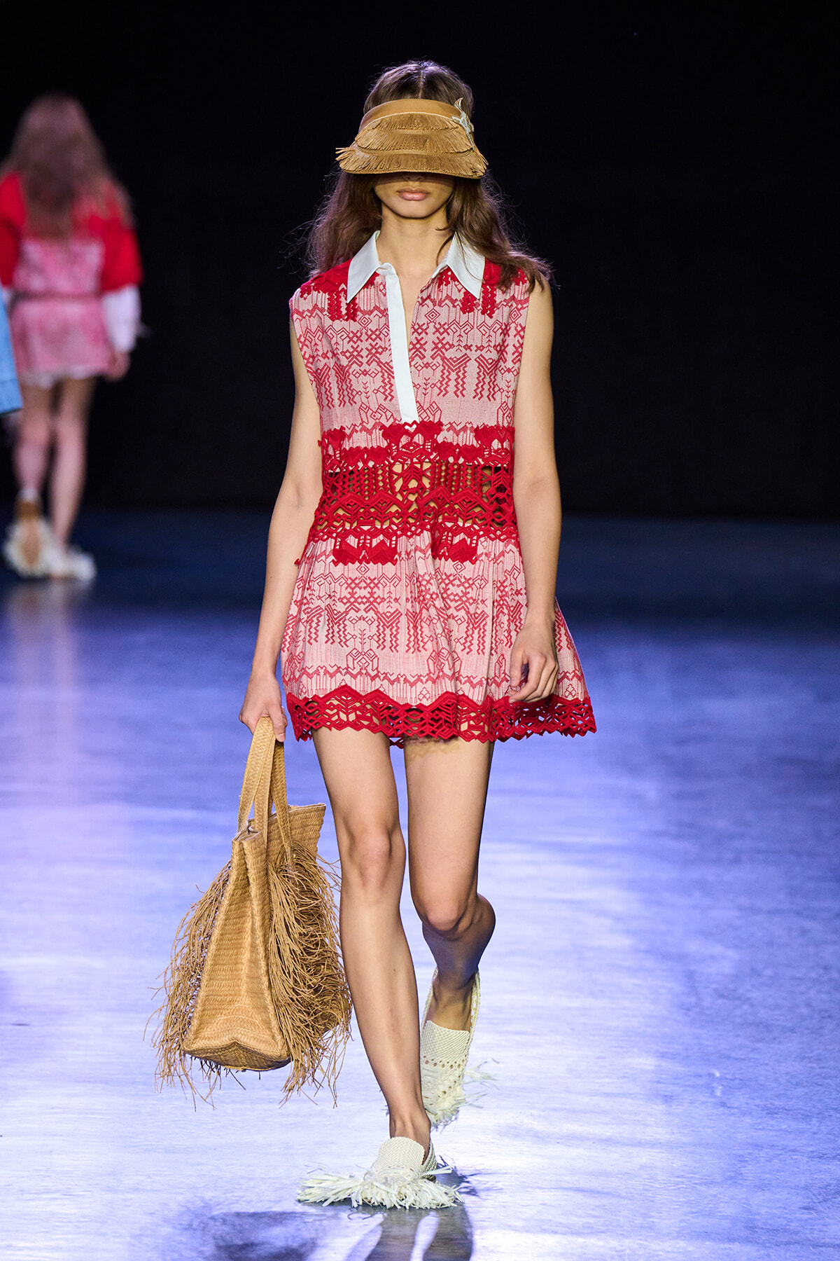 Runway model in pink and red lace dress with white collar, wearing a wide straw hat that covers her eyes, carrying a woven straw tote.