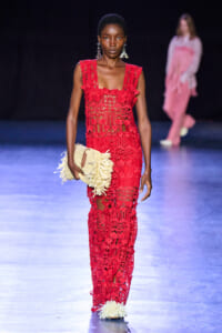 Model walks a fashion runway in a red lace gown, holding a cream fringed clutch; dark backdrop with another model blurred in pink in the background.