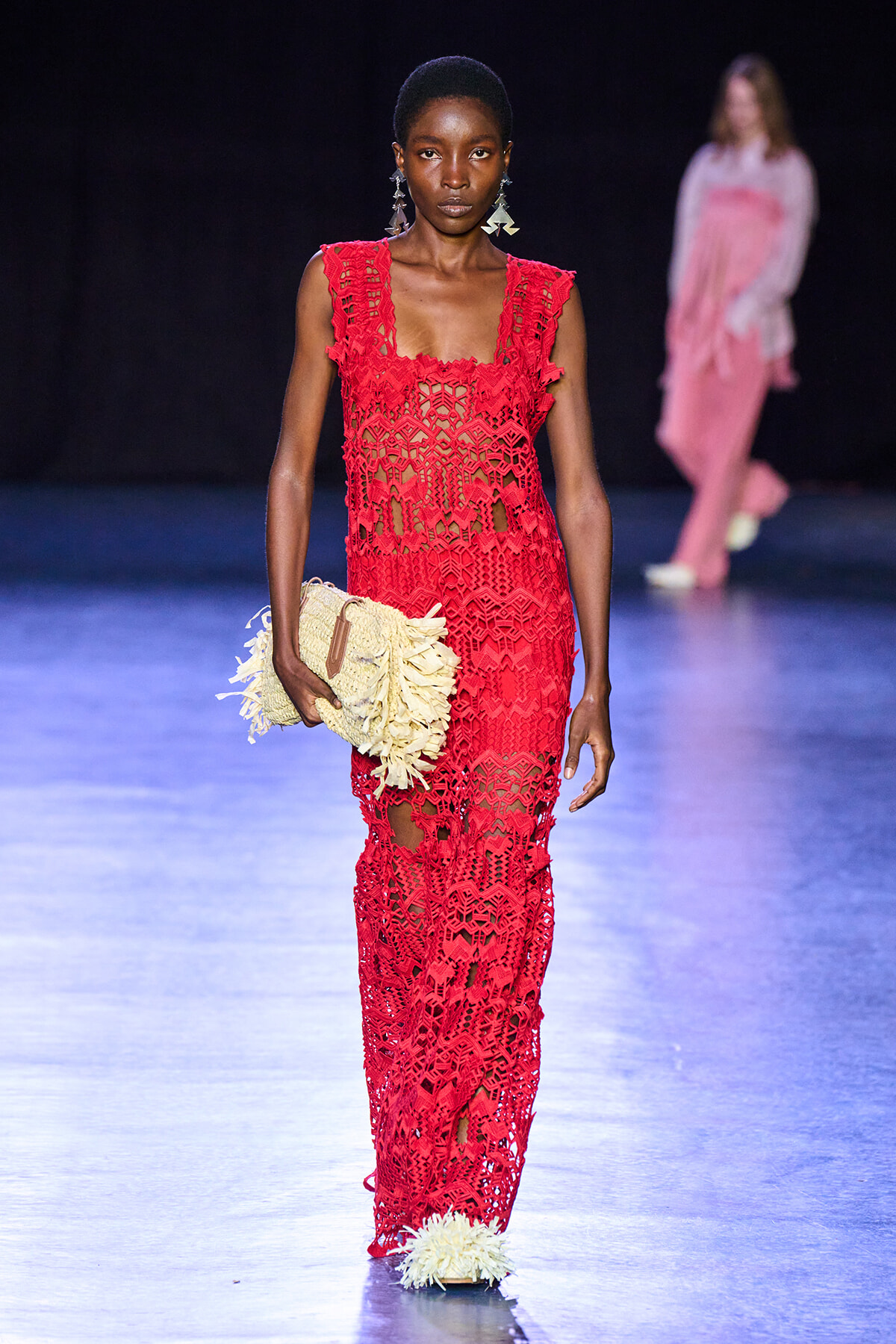 Model walks a fashion runway in a red lace gown, holding a cream fringed clutch; dark backdrop with another model blurred in pink in the background.