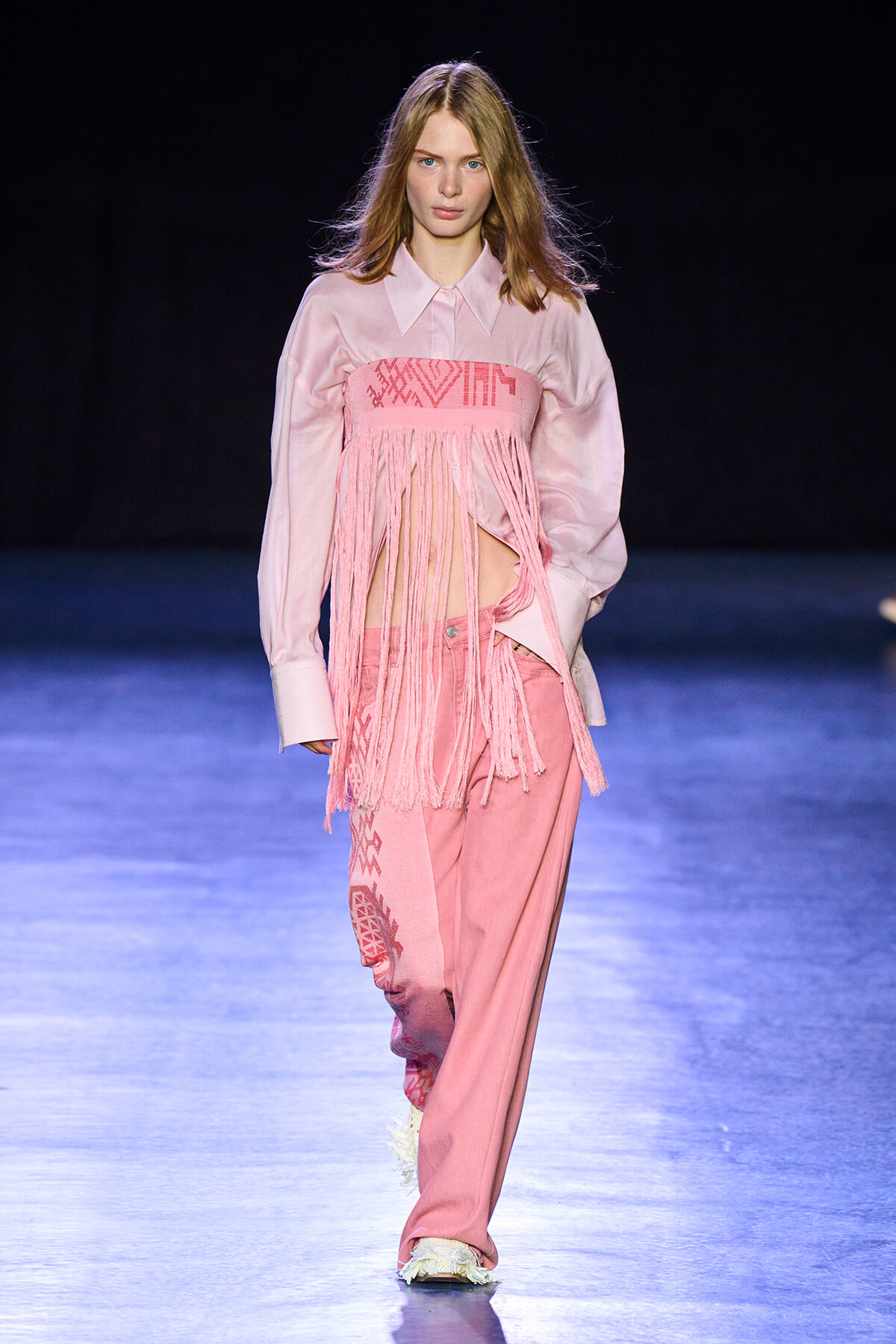 Model walks the runway in a pale pink button-down shirt with a pink fringe bandeau and matching pink pants on a blue-lit catwalk.