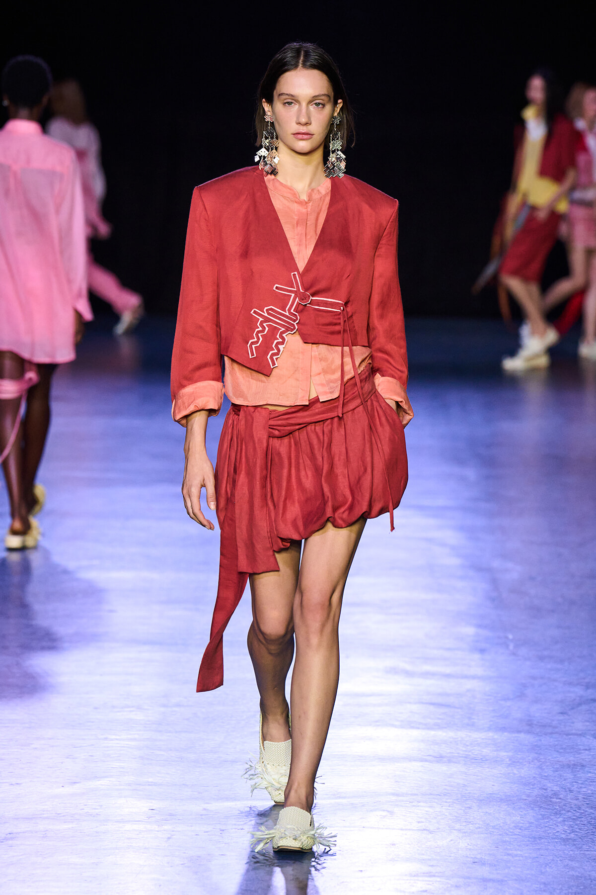 Model walking a fashion runway in a red layered ensemble with a pink underlayer and unique white stitched detailing on the top; oversized statement earrings complement the look.