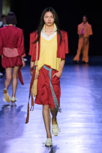 Model on a runway wearing a yellow knit vest over a white shirt, red blazer and red shorts, carrying a fringed crossbody bag.