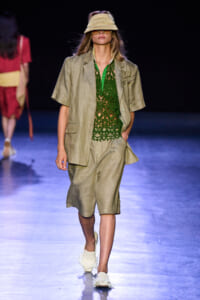 Model walking runway in beige suit with green lace top, straw visor, and white sneakers against dark backdrop.